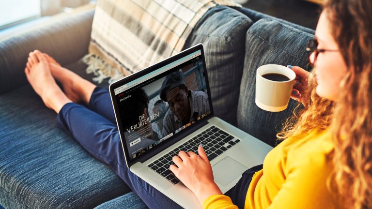 Eine Person liegt auf dem Sofa mit einer Tasse Kaffee in der Hand. Sie bedient ein MacBook, auf dem Netflix zu sehen ist.