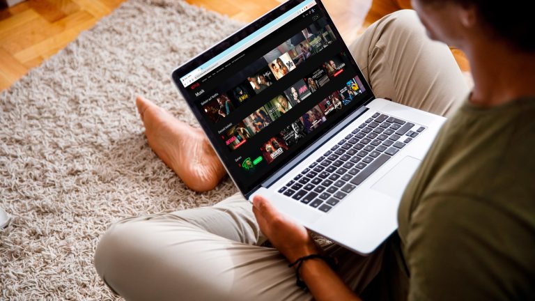Eine Person sitzt auf dem Boden mit einem MacBook auf dem Schoß. Auf dem Display ist Netflix zu sehen.