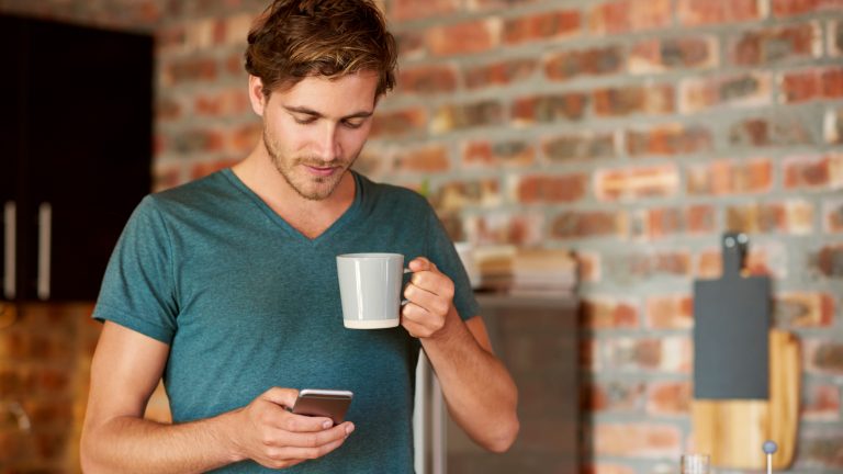 Eine Person trinkt eine Tasse Kaffee und bedient dabei ein Smartphone.