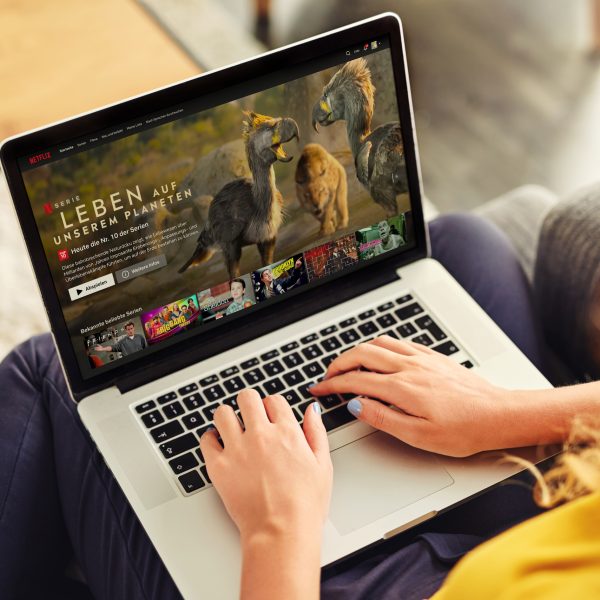 Eine Person sitzt mit einem Laptop auf dem Schoß auf einem Sofa. Auf dem Bildschirm ist die Netflix-Hauptseite zu sehen.