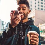 Eine Person l&auml;uft mit einem Kaffee in der Hand durch eine Stadt. Sie spricht dabei in ihr Smartphone.