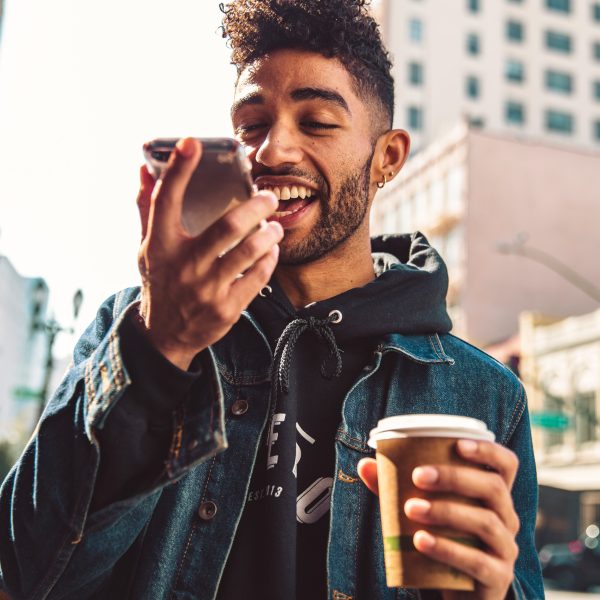Eine Person l&auml;uft mit einem Kaffee in der Hand durch eine Stadt. Sie spricht dabei in ihr Smartphone.