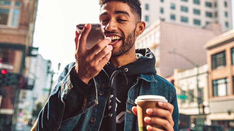 Eine Person l&auml;uft mit einem Kaffee in der Hand durch eine Stadt. Sie spricht dabei in ihr Smartphone.