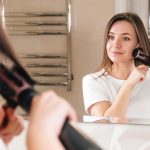 Person im wei&szlig;en T-Shirt steht vor Spiegel und gl&auml;ttet sich mit einer Warmluftb&uuml;rste die Haare.