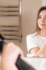 Person im weißen T-Shirt steht vor Spiegel und glättet sich mit einer Warmluftbürste die Haare.