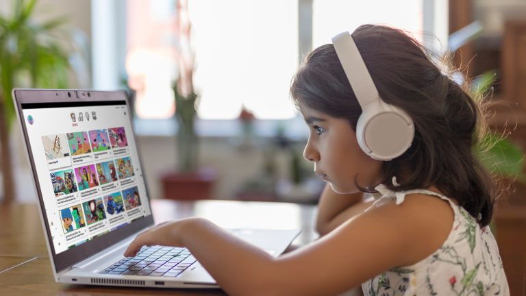 Junges Mädchen mit braunen langen Haaren sitzt mit weißen Kopfhörern vor einem Laptop mit YouTube-Inhalten.