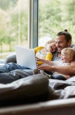 Ein Mann liegt mit seinen zwei jungen Kindern auf einer großen braunen Couch und sieht auf einem Laptop Videos.