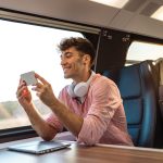 Eine Person mit Kopfh&ouml;rern um den Hals sitzt in einem Zug und schaut etwas auf ihrem Smartphone an.