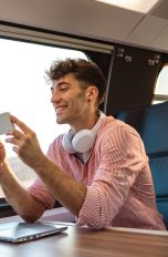 Eine Person mit Kopfhörern um den Hals sitzt in einem Zug und schaut etwas auf ihrem Smartphone an.