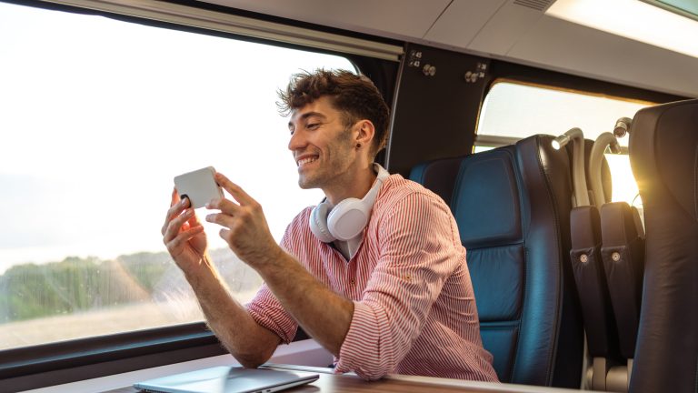 Eine Person mit Kopfhörern um den Hals sitzt in einem Zug und schaut etwas auf ihrem Smartphone an.