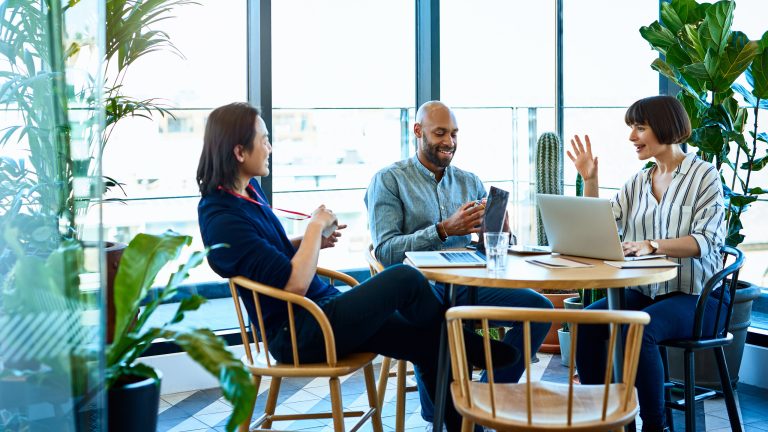 Drei Personen sitzen an einem runden Holztisch vor mehreren Laptops.