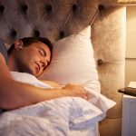 Eine Person liegt in Seitenlage im Bett. Im Ohr tr&auml;gt sie einen In-Ear-Kopfh&ouml;rer. Auf dem Nachttisch neben dem Bett liegt ein Smartphone.