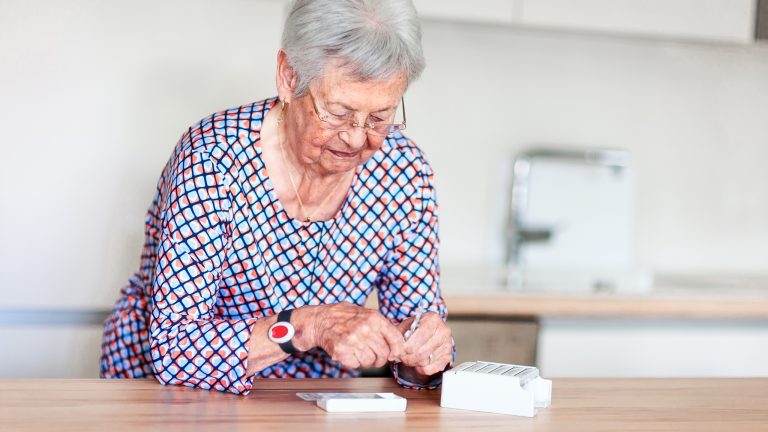 Eine ältere Person mit einem Hausnotruf-Knopf um das Handgelenk nimmt Tabletten.