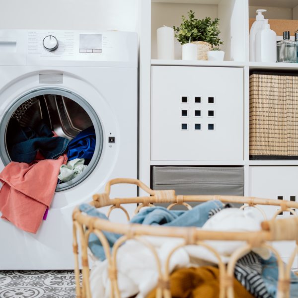 Ein Waschtrockner steht neben einem weißen Regal mit Deko. Im Vordergrund steht ein voller Wäschekorb.