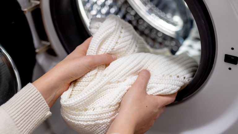 Zwei Hände holen einen weißen Wollpulli aus der Trommel einer Waschmaschine.