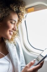 Eine Person mit voluminösen, lockigen Haaren sitzt am Fenster im Flugzeug und hört mit Kopfhörern Musik von ihrem Smartphone.