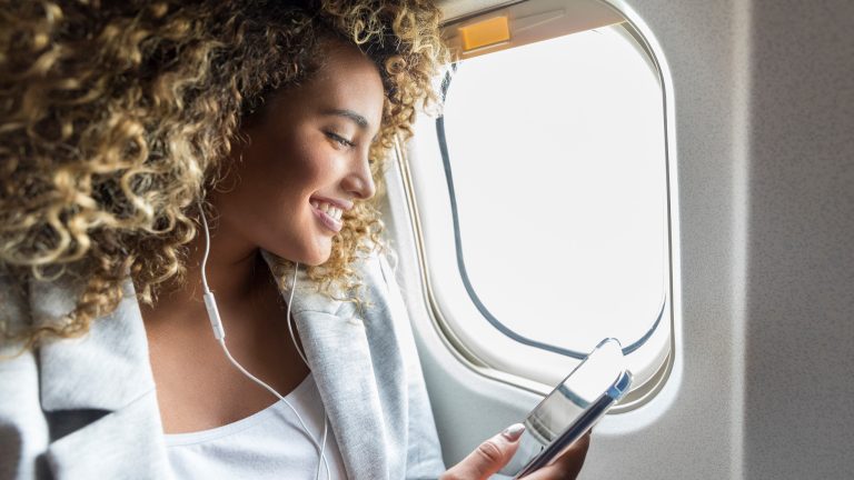 Eine Person mit voluminösen, lockigen Haaren sitzt am Fenster im Flugzeug und hört mit Kopfhörern Musik von ihrem Smartphone.