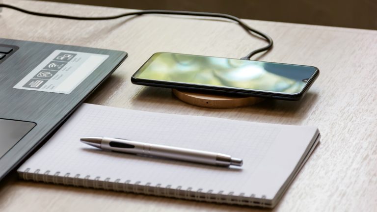Ein Smartphone liegt auf einem runden Wireless Charging Tool auf einem hellen Holztisch. Daneben liegt ein Notizbuch mit einem Kugelschreiber darauf. Im Anschnitt ist ein Laptop zu sehen.