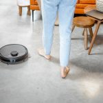 Ein schwarzer, kleiner, runder, flacher Wischroboter steht neben den F&uuml;&szlig;en einer Person in heller Jeans. Im Hintergrund stehen eine orange Couch und eine Couchtisch-Kombination aus Teakholz.