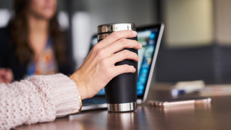Eine Person sitzt an einem REchner in einem B&uuml;ro und h&auml;lt eine Thermoskanne in der Hand.