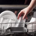 Eine Hand stellt ein Glas neben andere Gl&auml;ser und eine Sch&uuml;ssel in das obere Fach eines Geschirrsp&uuml;lers.