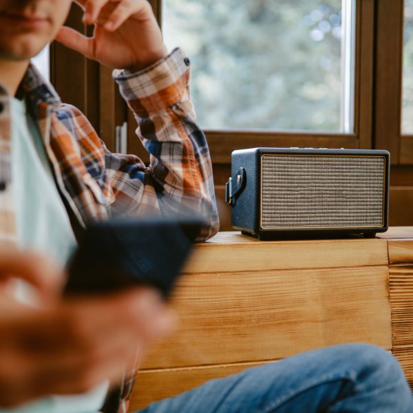 Eine Person mit Smartphone in der Hand lehnt an einer Holzkommode, auf der ein Bluetooth-Lautsprecher steht.