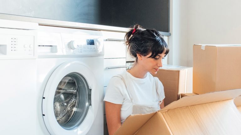 Eine Person sitzt vor einer Waschmaschine und schaut in Umzugskartons.