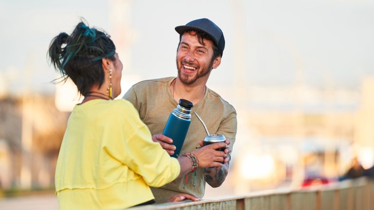 Zwei Personen stehen im Freien und lachen. Eine Person reicht der anderen eine Thermoskanne.