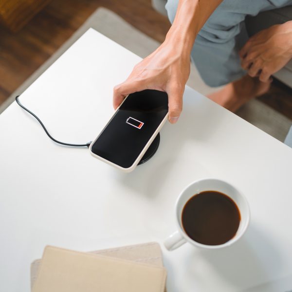 Eine Person in hellen Jeans legt ein Smartphone auf ein rundes wireless charging tool auf einem wei&szlig;en Tisch. Daneben steht eine Tasse Kaffee.