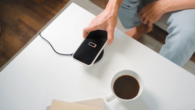 Eine Person in hellen Jeans legt ein Smartphone auf ein rundes wireless charging tool auf einem weißen Tisch. Daneben steht eine Tasse Kaffee.