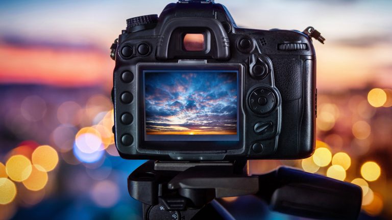 In der Mitte und im Fokus sieht man die Rückseite einer Spiegelreflexkamera. Auf dem Bildschirm sieht man einen Sonnenuntergang mit Wolken. Drumherum sind verschwommene Lichtpunkte zu sehen.
