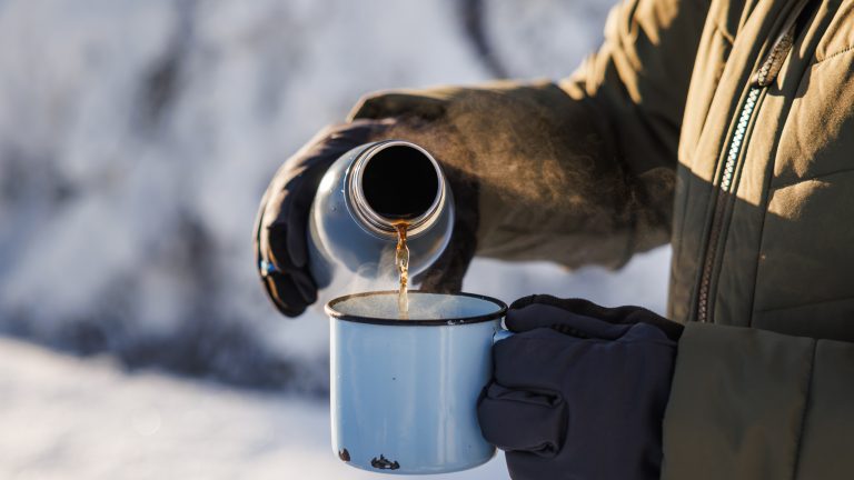 Eine Person in einer Schneelandschaft gie&szlig;t Fl&uuml;ssigkeit von einer Thermoskanne in eine Tasse.