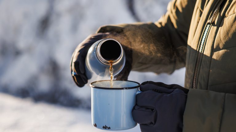 Eine Person in einer Schneelandschaft gießt Flüssigkeit von einer Thermoskanne in eine Tasse.