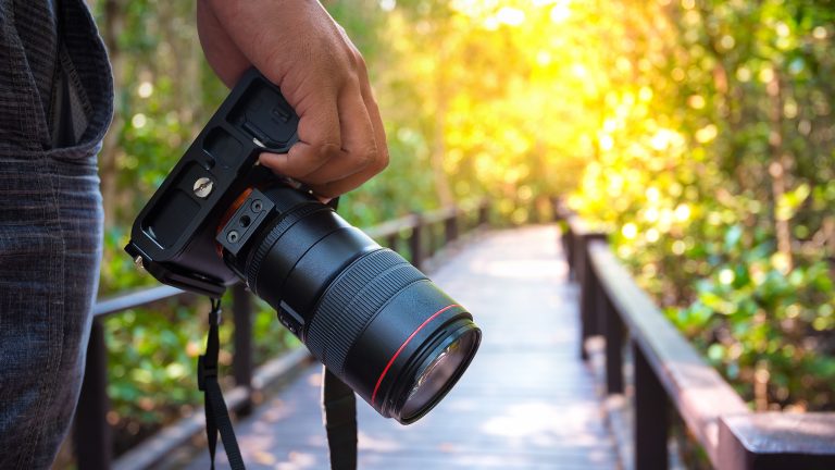 Eine Hand hält eine Kamera mit mittelgroßem Objektiv nach unten. Im Hintergrund sieht man einen Holzweg mit Holzgeländer in einem Wald.