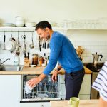 Eine Person stellt l&auml;chelnd Tassen in einen Geschirrsp&uuml;ler, eine weitere Person schaut zu.
