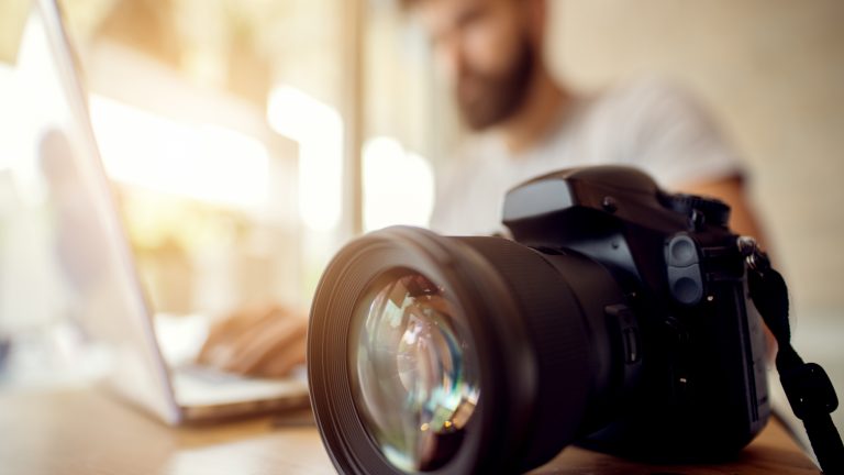 Eine Kamera liegt auf einem Holztisch. Im Hintergrund sieht man verschwommen eine Person mit Bart an einem Laptop.