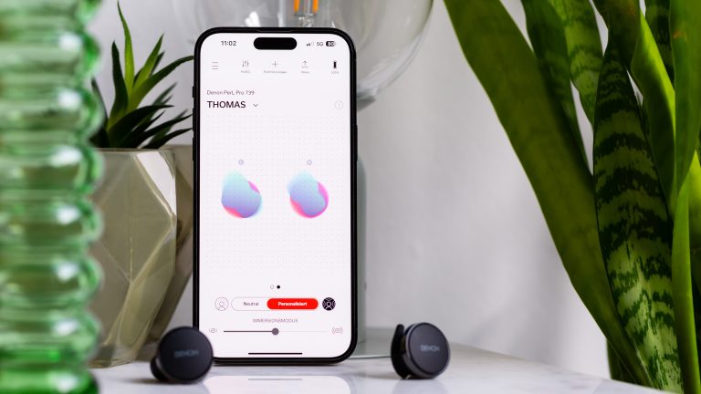 Ein iPhone mit geöffneter Denon-App steht auf einem Tisch. Davor liegen die PerL Pro.