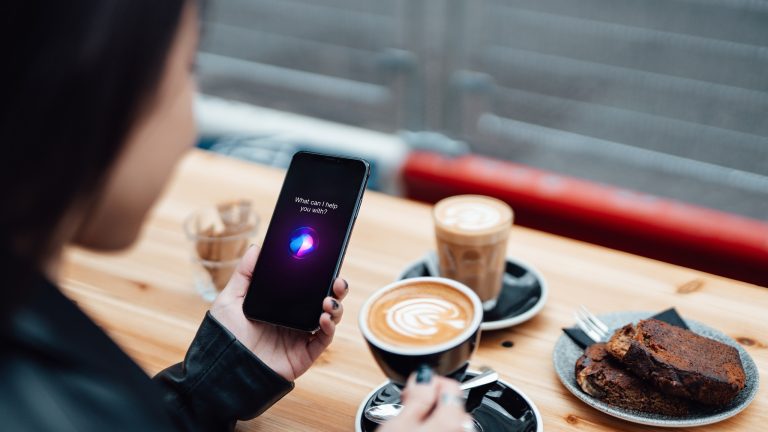 Eine Person in einem Café hält ein Smartphone in der Hand, auf dem eine Sprachassistenz gerade zuhört. In der anderen Hand hält sie eine Kaffeetasse.