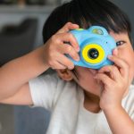 Ein Kind schaut durch den Sucher einer Kinderkamera Richtung Betrachter*in.