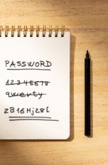 Notizblock auf einem Holztisch mit dem Begriff ‘PASSWORD’ oben und einer Liste von einfachen, durchgestrichenen Passwörtern wie ‘1234567’. Das letzte Passwort ist komplex und doppelt unterstrichen. Neben dem Notizbuch liegt ein schwarzer Stift.