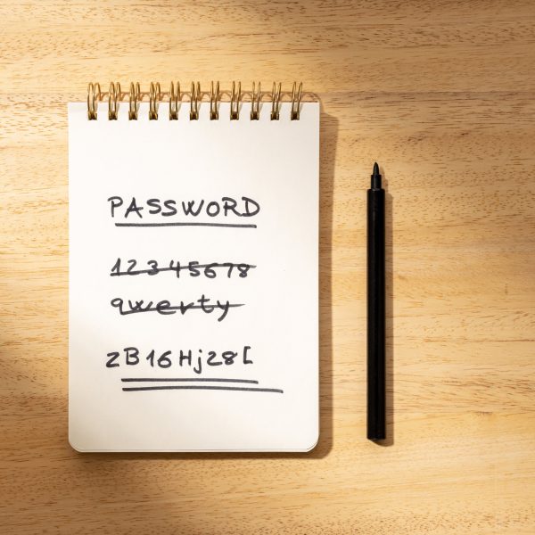 Notizblock auf einem Holztisch mit dem Begriff ‘PASSWORD’ oben und einer Liste von einfachen, durchgestrichenen Passwörtern wie ‘1234567’. Das letzte Passwort ist komplex und doppelt unterstrichen. Neben dem Notizbuch liegt ein schwarzer Stift.