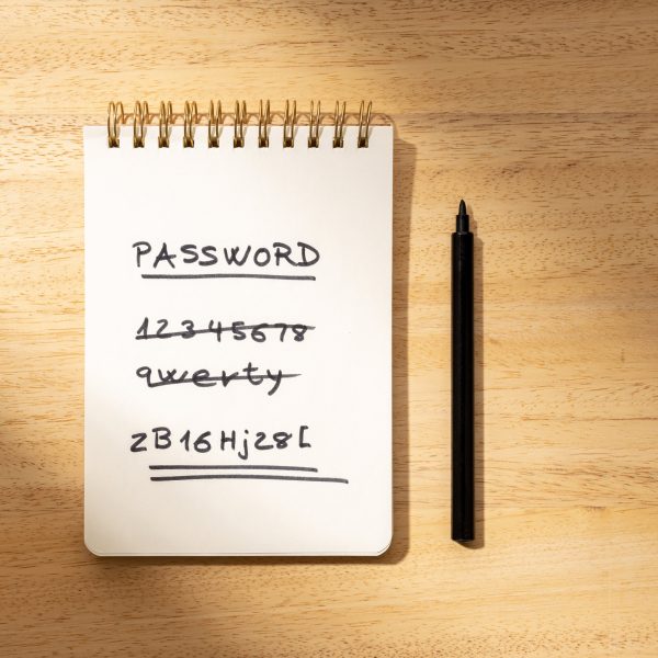 Notizblock auf einem Holztisch mit dem Begriff &lsquo;PASSWORD&rsquo; oben und einer Liste von einfachen, durchgestrichenen Passw&ouml;rtern wie &lsquo;1234567&rsquo;. Das letzte Passwort ist komplex und doppelt unterstrichen. Neben dem Notizbuch liegt ein schwarzer Stift.
