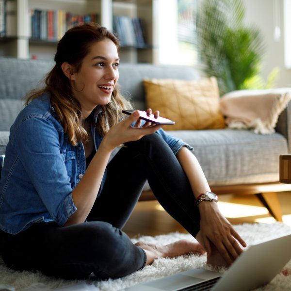 Eine Person sitzt auf einem Teppich vor einem Sofa. In der Hand hält sie ein Smartphone flach vor den Mund. Vor ihr steht ein aufgeklappter Laptop.