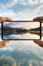 Ich-Perspektive einer Person, die ein Smartphone auf eine Seelandschaft richtet und ein Foto davon macht.