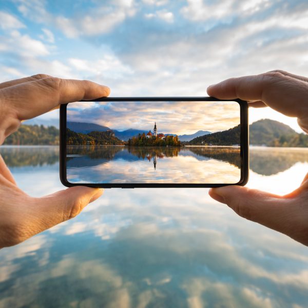 Ich-Perspektive einer Person, die ein Smartphone auf eine Seelandschaft richtet und ein Foto davon macht.