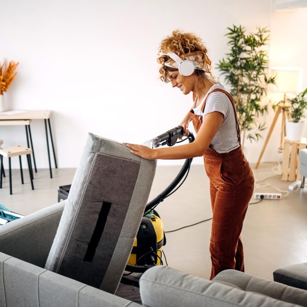 Eine Person mit blonden Locken und wei&szlig;en Kopfh&ouml;rern reinigt mit einem schwarz gelben Nasssauger die Polster einer grauen Couch. Im Hintergrund sieht man einen kleinen Schreibtisch und Pflanzen.