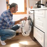 Eine Person sitzt in der Hocke vor einer Waschmaschine und h&auml;lt die T&uuml;r auf.