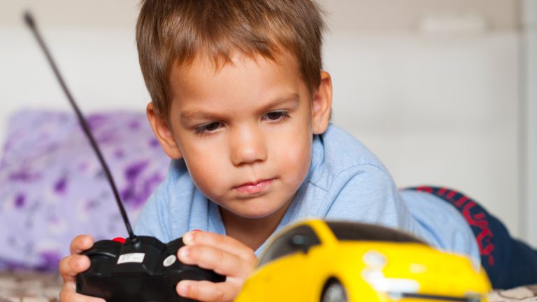 Ferngesteuerte Autos für Kinder: Darauf kommt es an Ein Kind liegt auf einem Fußboden und steuert einen gelben ferngesteuerten Fiat 500 mit einer Fernbedienung.