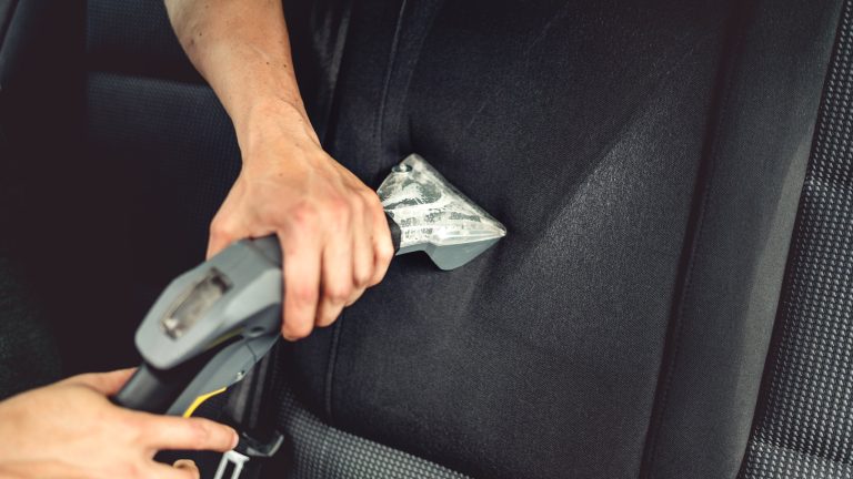 Zwei kräftige Hände halten die graue Polsterdüse eines Nasssaugers an einen Autositz und reinigen diesen damit.