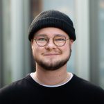 Junger Mann mit runder Brille, schwarzer Wollmütze, Bart und schwarzem Pullover.